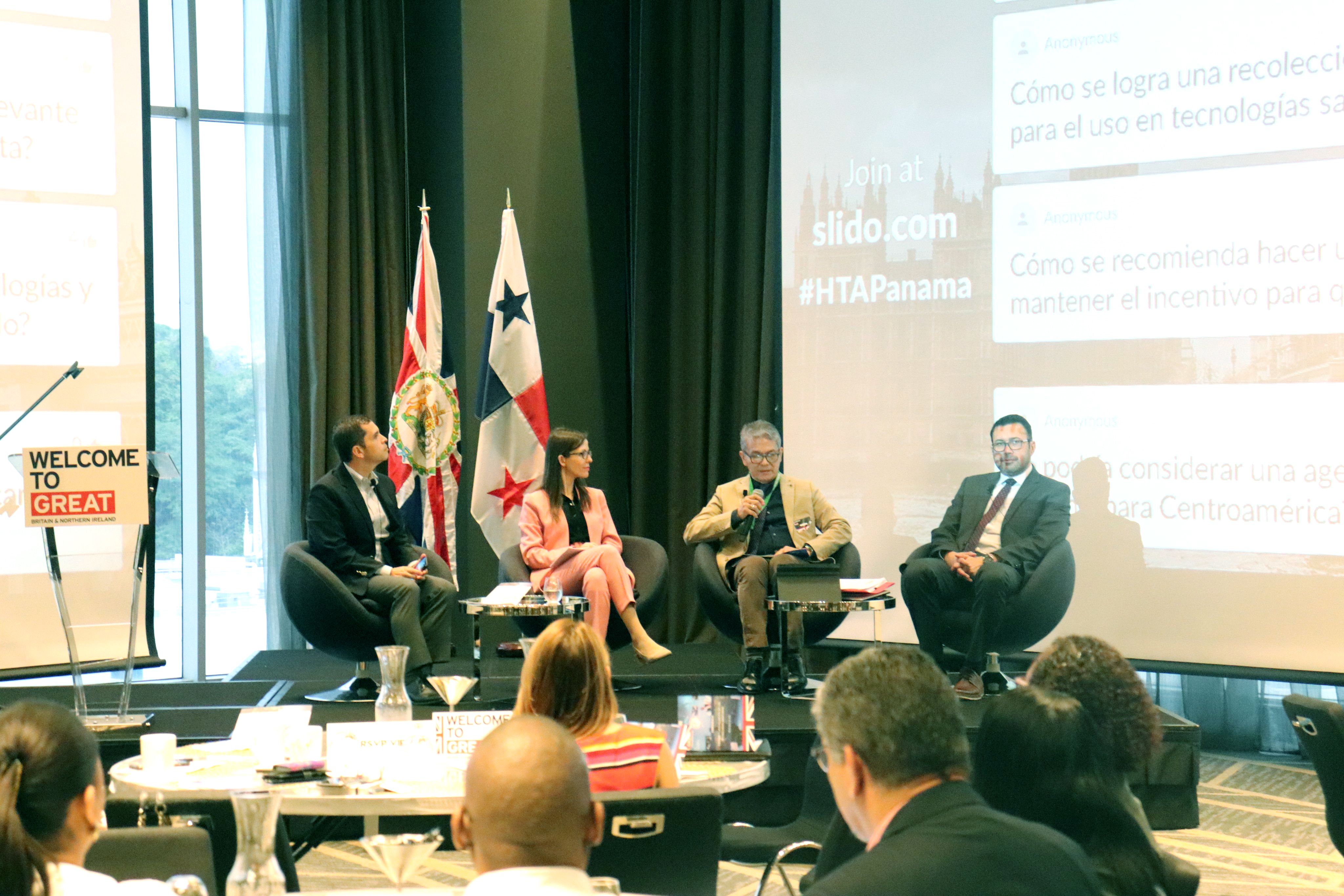 Four people sitting in chairs on a stage, presenting at a conference. They are in front of a large screen with a presentation playing on it. You can see the heads of the audience, who are seated round tables, just in front of them.